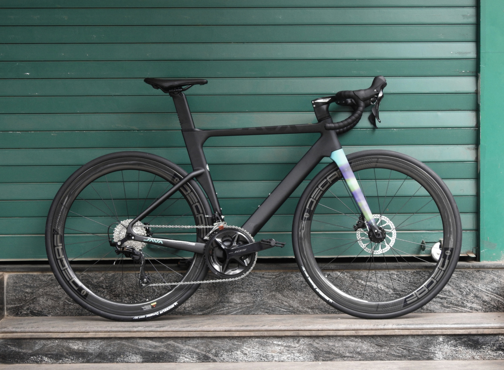A matte black carbon fiber road bicycle with drop handlebars and deep wheels, parked against a dark green corrugated metal wall.