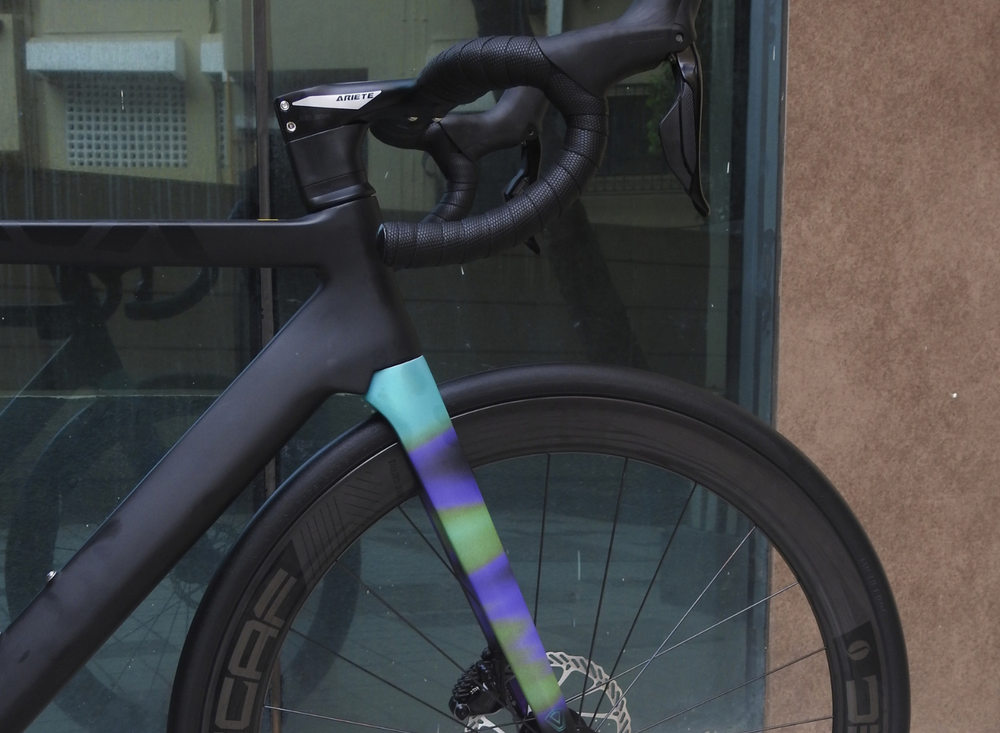 Close-up of a black road bicycle frame and front wheel leaning against a glass door, with a colorful gradient decal running from the handlebar area down the front triangle and fork.