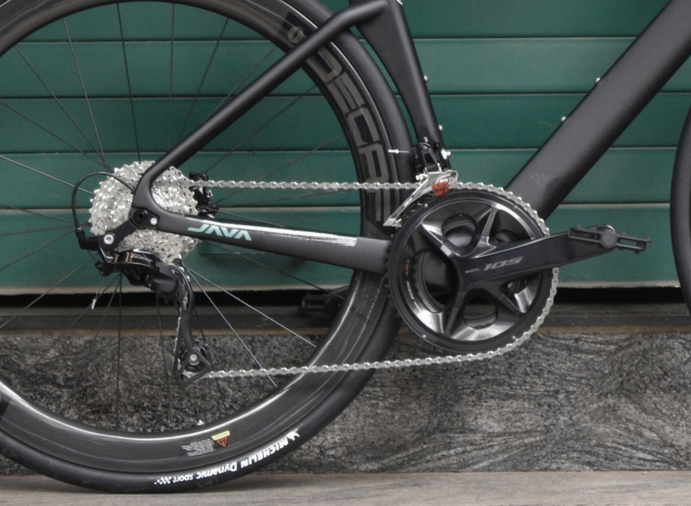 Close-up of a black road bicycle's rear wheel, chainring, cassette, and frame with visible Java and SRAM branding, set against a green paneled wall.