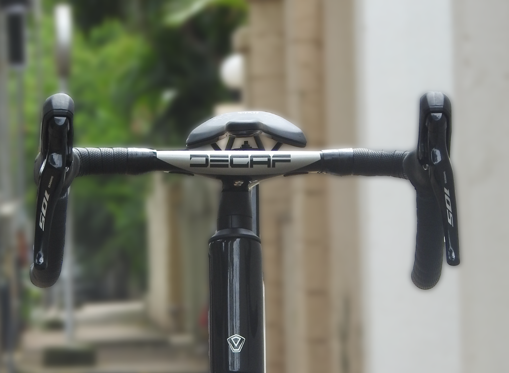 Close-up of a bicycle drop handlebar with black handlebar tape, black brake levers, and a silver Decaf center section, set against a blurred outdoor background.