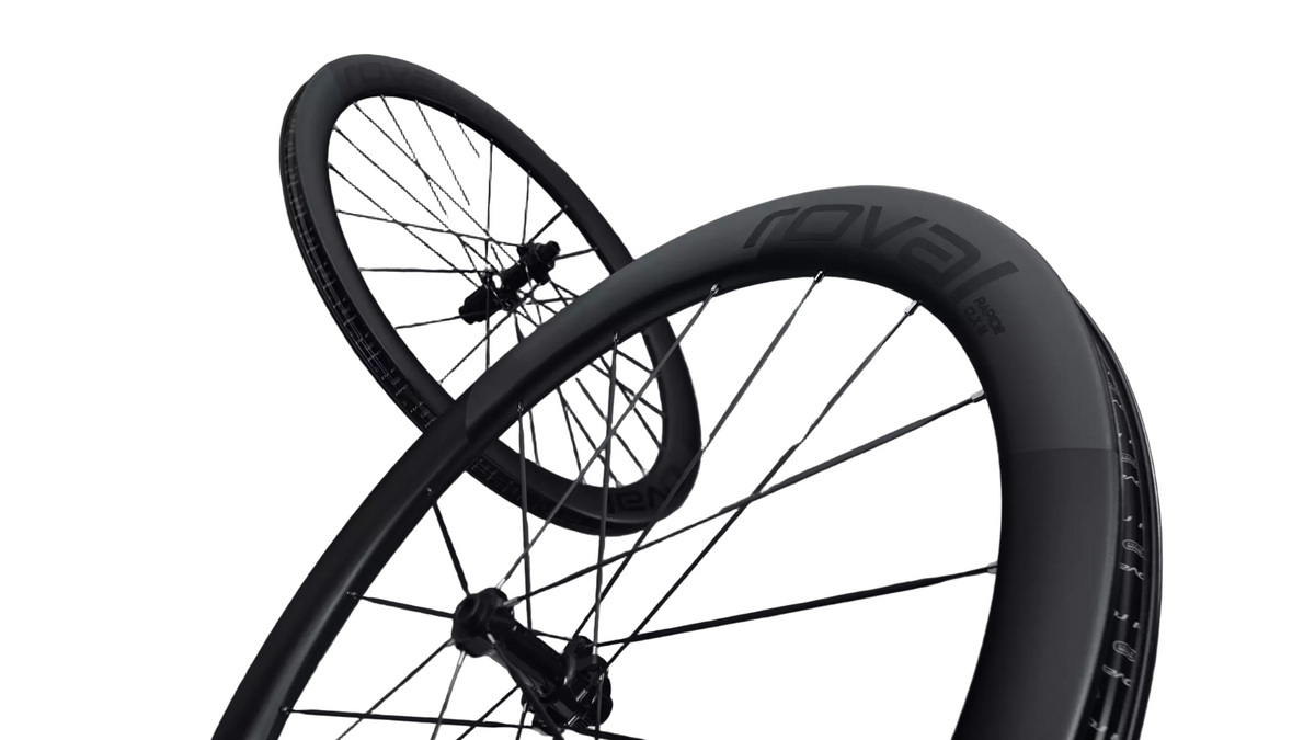 Two black bicycle wheels with thin spokes and dark rims, one angled behind the other against a dark gray studio background. The front wheel rim shows the brand name 'Royal' in dark lettering.